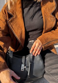 Hnědá semišová bunda přes černý top, držící černou koženou tašku se stříbrným kováním, která má zip a krátkou rukojeť.