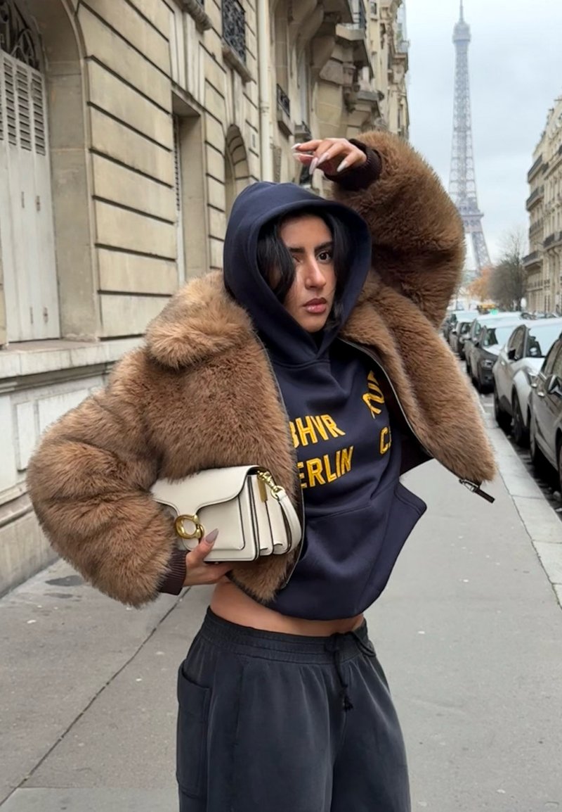 Faux fur brown bomber jacket with a hood, navy sweatshirt featuring yellow text, black joggers, and a small white handbag with gold accents.