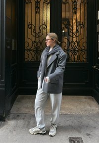 Veste blazer grise oversize, pull gris clair et pantalon de survêtement. Accessoires comprenant une écharpe sombre et des baskets beiges. Porte vintage en arrière-plan.