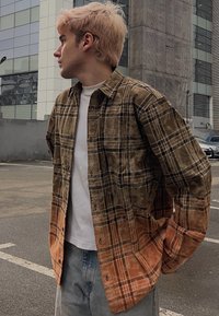 Jeune homme aux cheveux blonds portant une chemise à carreaux marron et orange, un t-shirt blanc et un jean, se tenant dans un parking urbain.