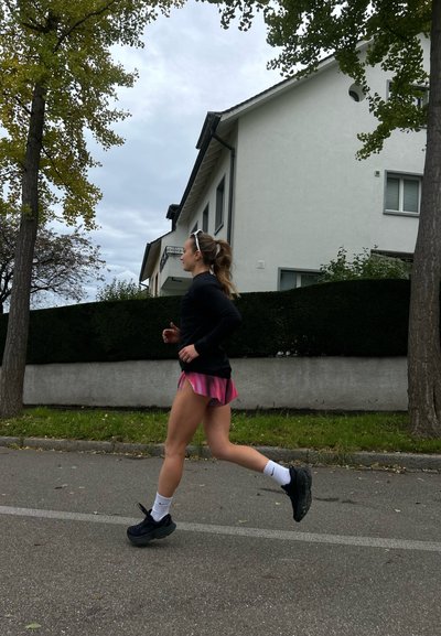 Mujer atlética corriendo por un camino, vestida con una camiseta de manga larga negra, pantalones cortos rosas, calcetines blancos y zapatos negros. Árboles y una casa al fondo.