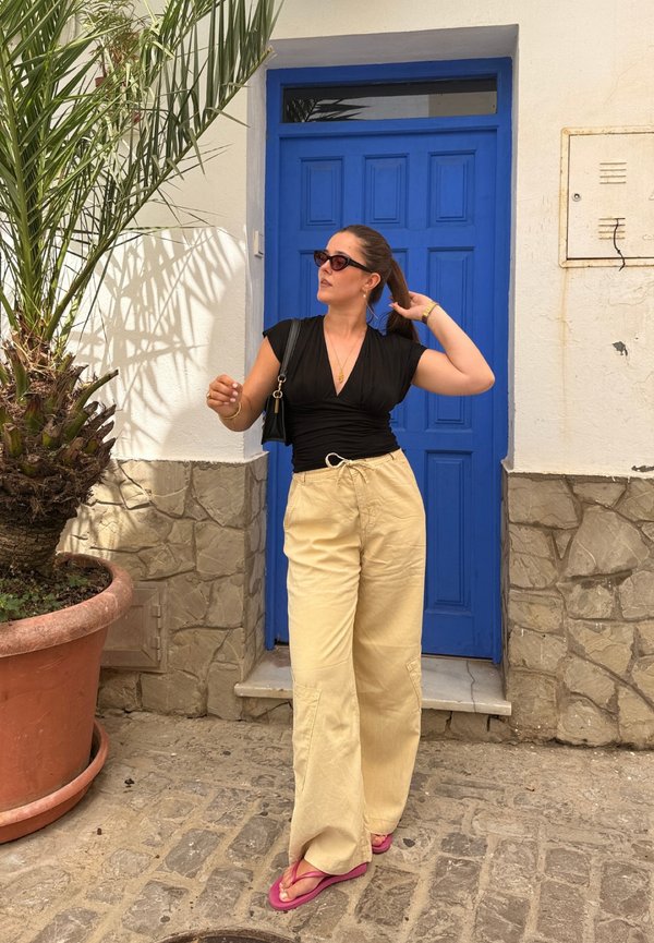 Femme en pantalon beige, haut noir et sandales roses, debout dans une rue pavée, ajustant ses cheveux près d'une porte bleu vif et d'un grand palmier en pot.