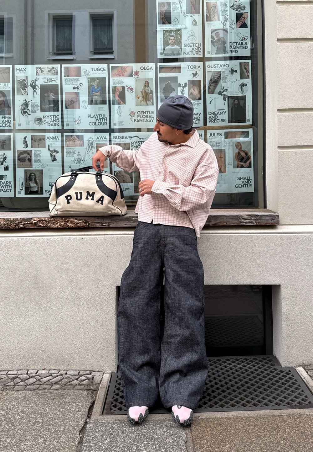 Witte duffeltas met zwarte accenten en "PUMA"-logo, lichtroze geruit overhemd, oversized donkere denim broek, grijze muts en roze schoeisel.