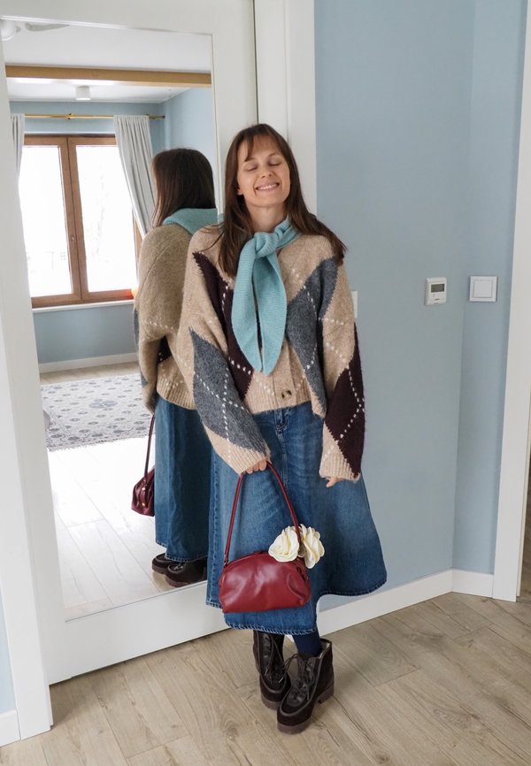 Junge Frau lächelt mit geschlossenen Augen, trägt einen gemusterten Pullover, einen türkisfarbenen Schal, einen Jeansrock, schwarze Stiefel und hält eine rote Handtasche mit weißen Blumen in Innenräumen.