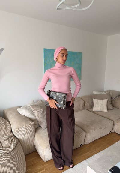 Suéter de cuello alto de canalé rosa claro, pantalones anchos marrones de pierna ancha y un bolso de mano negro con tachuelas. Estilizado en un entorno moderno interior.