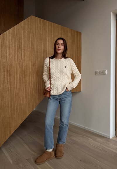 Suéter blanco de punto trenzado con puños acanalados, jeans azules y botas de tobillo color marrón. Bolso de hombro marrón. Fondo de panel de madera texturizada.