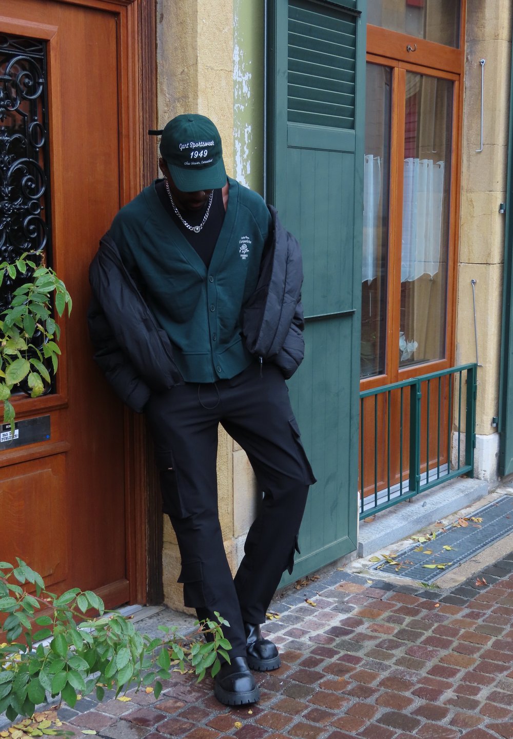 Giacca a vento verde indossata sopra un cardigan verde e una maglietta nera, abbinata a pantaloni cargo neri e stivali neri massicci.
