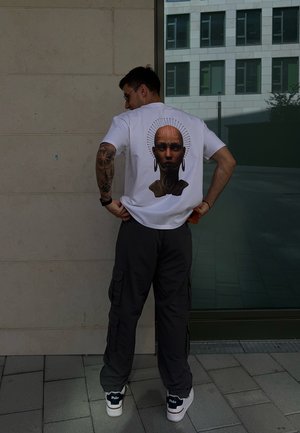 Camiseta blanca con un gráfico de una cabeza calva con pendientes y un halo decorativo, combinada con pantalones cargo oscuros y zapatillas blancas.