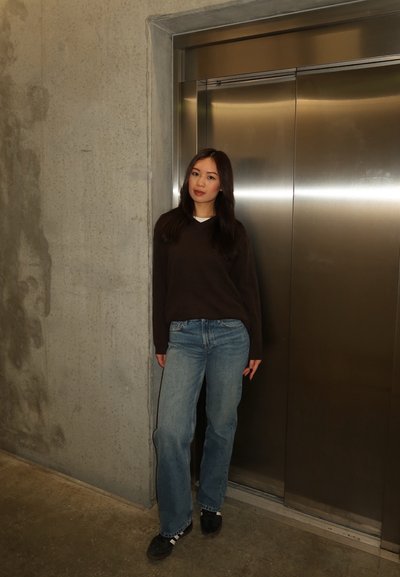 Joven mujer con suéter oscuro y jeans, de pie frente a puertas metálicas cerradas de un ascensor en un pasillo de concreto.