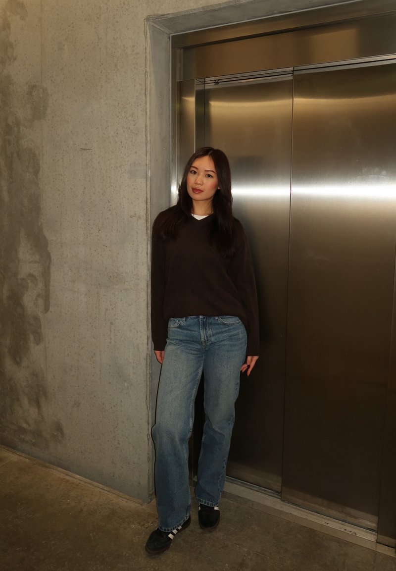 Jeune femme portant un pull sombre et un jean, debout devant des portes d'ascenseur métalliques fermées dans un couloir en béton.