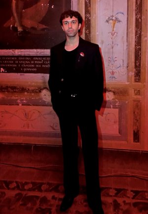 Costume noir ajusté avec une coupe slim, associé à une chemise noire. L'arrière-plan présente une décoration murale ornée et fanée dans des couleurs atténuées.