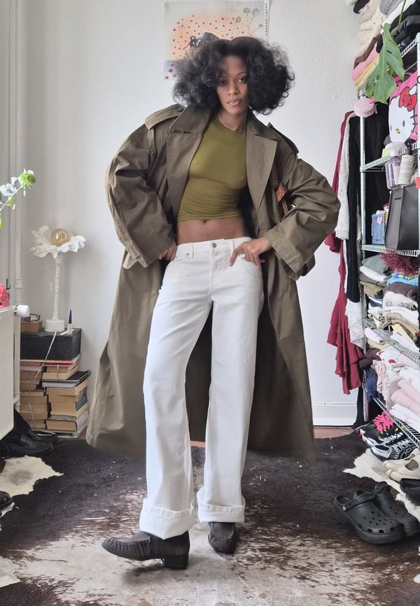 Woman with curly hair wearing an olive green crop top, long brown coat, white pants, and dark shoes standing in a room with books and clothes.