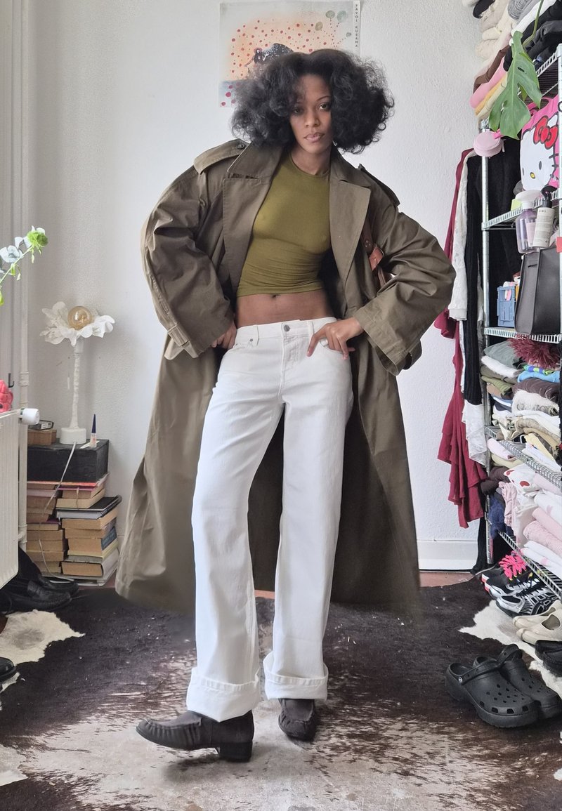 Kvinna med lockigt hår som bär en olivgrön crop top, en lång brun kappa, vita byxor och mörka skor, stående i ett rum med böcker och kläder.