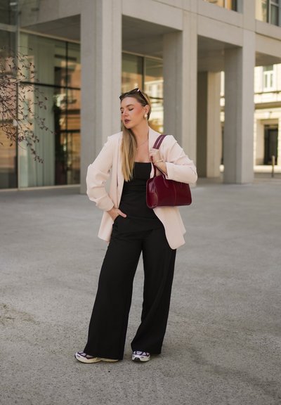 Blazer rosa claro sobre una blusa negra, combinado con pantalones anchos negros y zapatillas blancas. Lleva un bolso burdeos. Texturas suaves, diseño moderno.