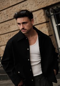 Svart knappet jakke med avslappet passform, sølvdetaljer og lommer, båret over en hvit singlet. Lys, teksturert bakgrunn.