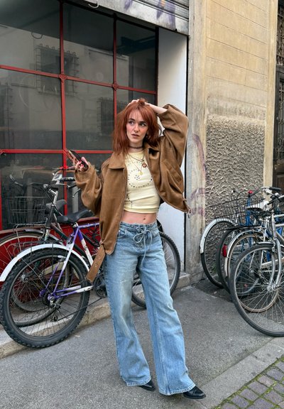 Mujer joven con cabello rojo, vestida con chaqueta marrón y jeans, de pie junto a bicicletas estacionadas en la acera de la ciudad, sosteniendo un teléfono y tocándose el cabello.