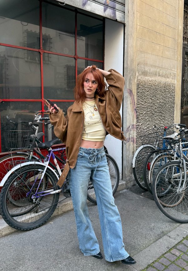 Mladá žena s červenými vlasmi, oblečená v hnedej bunde a rifle, stojí pri zaparkovaných bicykloch na mestskom chodníku, drží telefón a dotýka sa vlasov.