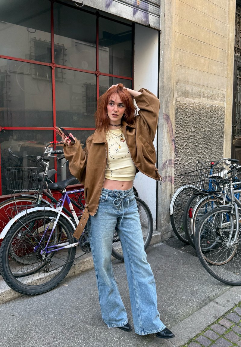 Jeune femme aux cheveux roux portant une veste marron et un jean, debout près de vélos stationnés sur un trottoir urbain, tenant un téléphone et se touchant les cheveux.