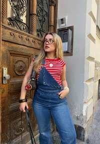 Denim dungarees with scalloped edges, worn over a red and white striped top. Accessories include a small black bag and decorative bracelets.
