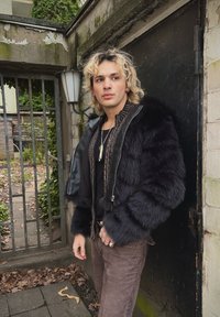 Μαύρο ψεύτικο γούνα μπουφάν με δερμάτινες λεπτομέρειες, ραμμένο κολάρο και κλείσιμο με φερμουάρ. Φορεμένο πάνω από μαύρη μπλούζα, συνδυασμένο με καφέ παντελόνι. Αστική εξωτερική τοποθεσία.