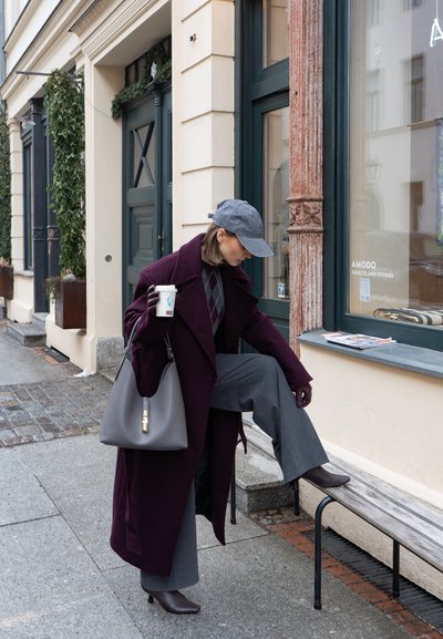 Mujer con abrigo morado y gorra gris sosteniendo una taza de café y ajustándose la bota del tobillo mientras está de pie junto a un banco frente a una tienda.