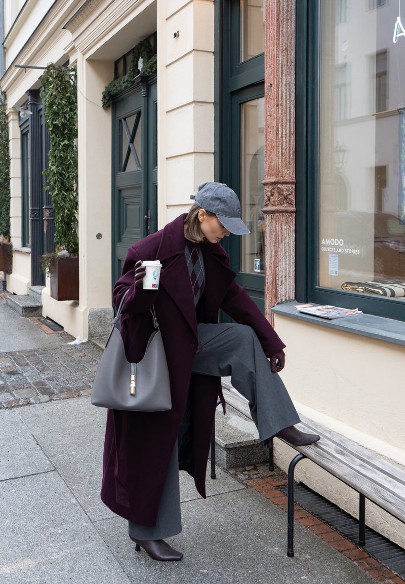 Femme en manteau violet et casquette grise tenant une tasse de café et ajustant sa bottine tout en se tenant près d'un banc devant une vitrine.