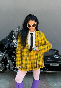 Casaco de fato xadrez amarelo com uma camisa branca e uma gravata preta; botas roxas até ao joelho; óculos de sol brancos oversized; collants com padrão floral.