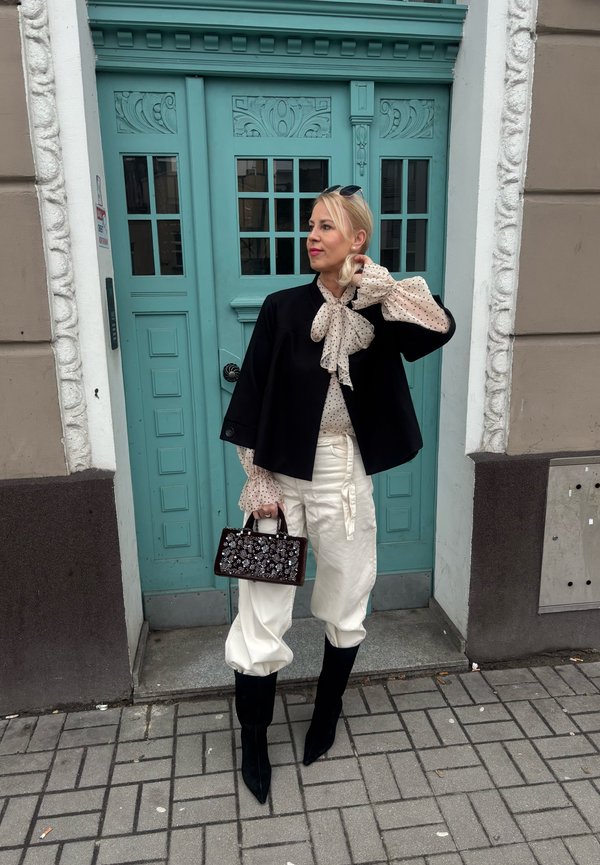 Vrouw in een zwart jasje, polkadot-blouse met strik, witte broek, zwarte laarzen, met een versierde handtas, staand voor blauwgroene decoratieve dubbele deuren.