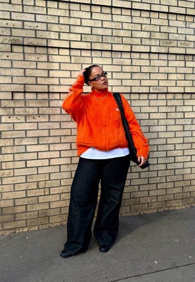 Persona con gafas, suéter peludo de color naranja brillante, camisa blanca, pantalones negros de pierna ancha, llevando una bolsa negra, de pie frente a una pared de ladrillos beige.