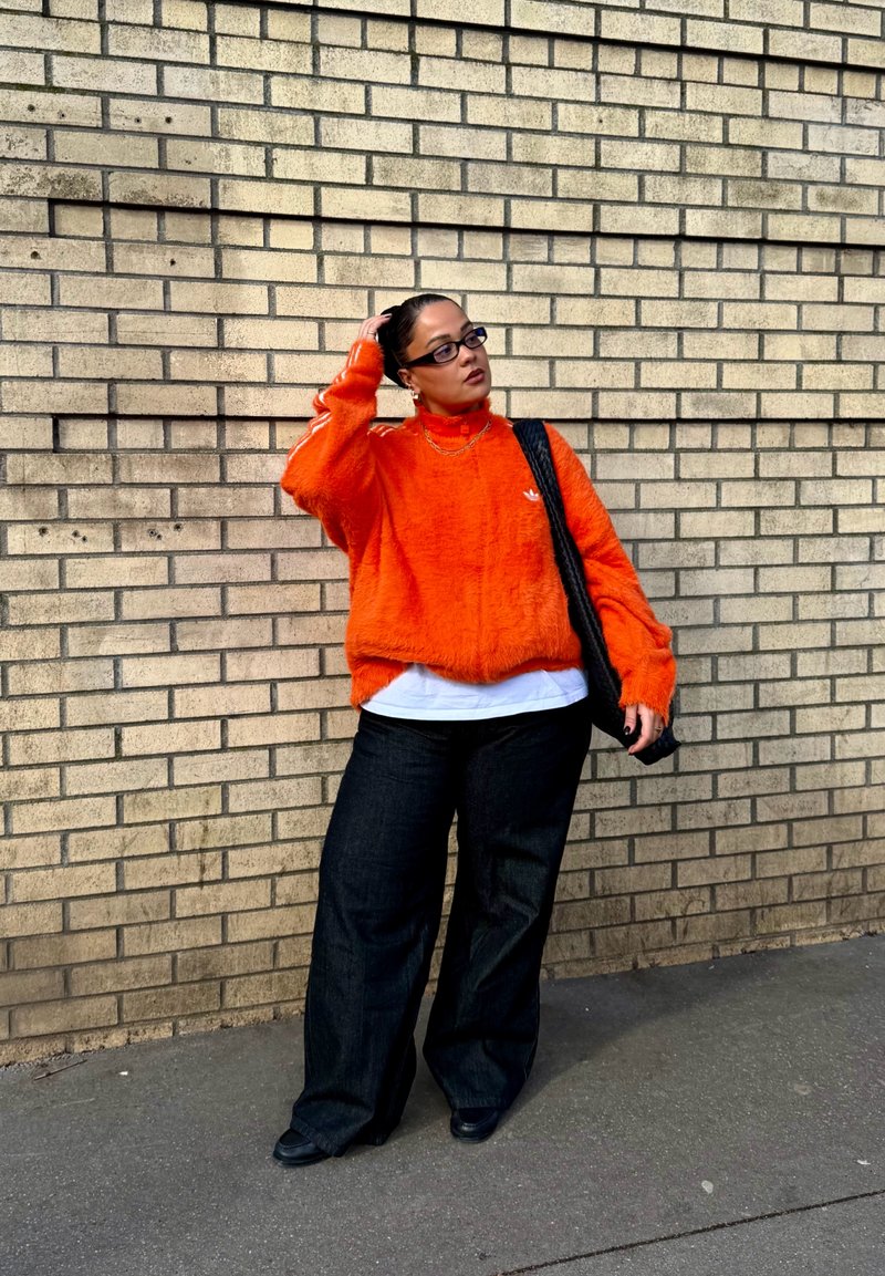 Person mit Brille, trägt einen leuchtend orangefarbenen, kuscheligen Pullover, weißes Hemd, schwarze Hosen mit weitem Bein, hält eine schwarze Tasche, steht vor einer beigen Ziegelwand.