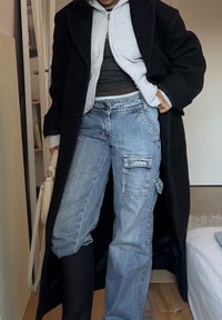 Manteau long noir, sweat à capuche gris zippé, débardeur foncé, jean cargo bleu clair avec poches, bottines noires, tissu texturé.