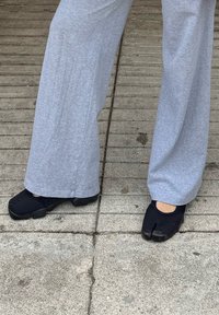 Fekete, rugalmas anyagból készült lábbeli, osztott orr-résszel, szürke, bő szárú nadrággal viselve, texturált betonfelületen.