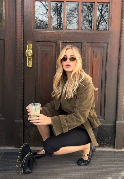 Mujer rubia con gafas de sol, chaqueta marrón y pantalones negros agachada junto a una puerta de madera, sosteniendo una bebida verde con una bolsa negra cerca.