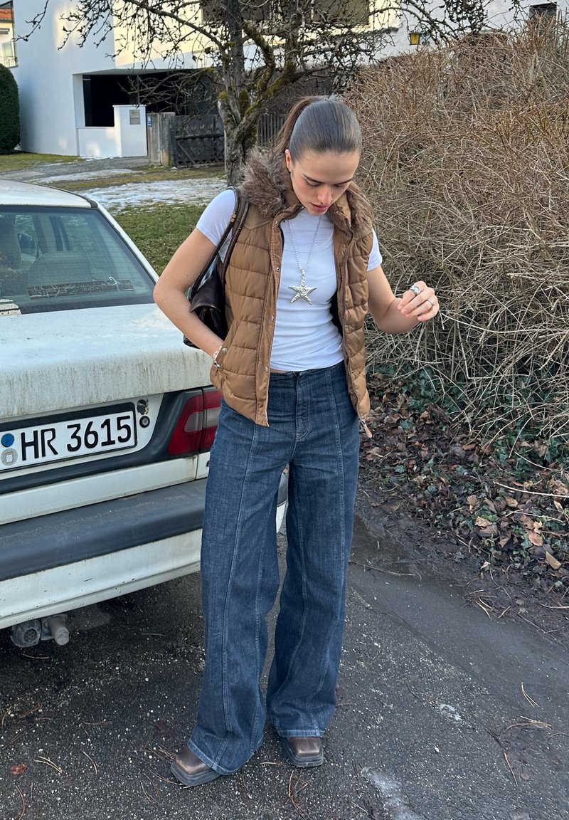 Young woman wearing a brown fur-hooded gilet, white t-shirt, and wide-leg jeans stands near a parked white car on a suburban street.