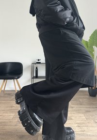 Schwarze, weit geschnittene Hosen aus glattem Stoff, kombiniert mit klobigen schwarzen Plateau-Schuhen mit auffälligen Metalldetails.