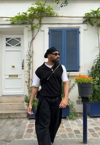 Μαύρο μανίκι χωρίς ρεβέρ πάνω από λευκό πουκάμισο, φαρδιά μαύρα παντελόνια, μαύρο μπερέ και γυαλιά ηλίου. Το φόντο διαθέτει μια λευκή πόρτα και μπλε παραθυρόφυλλα.
