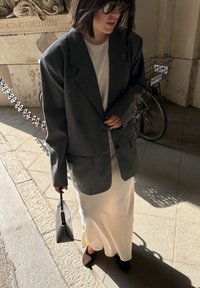 Overmatig grijze blazer over een witte jurk, gecombineerd met zwarte schoenen. In één hand wordt een kleine zwarte handtas vastgehouden, terwijl er op stenen straatstenen wordt gestaan.