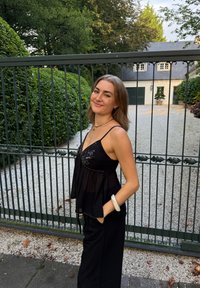 Top senza maniche nero con sovrapposizione trasparente e decorazioni di perline, abbinato a pantaloni neri. Sullo sfondo si possono vedere delle piante e un cancello.