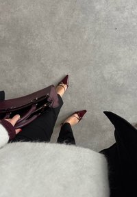 Personne debout sur un sol gris portant des ballerines pointues bordeaux, un pantalon noir, un pull blanc duveteux, tenant un sac à main assorti bordeaux.