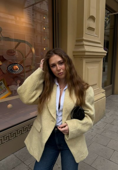 Joven mujer con cabello largo y ondulado, lleva un abrigo cremoso y peludo, camisa blanca y vaqueros azules, está de pie en la acera de la ciudad cerca de un escaparate.