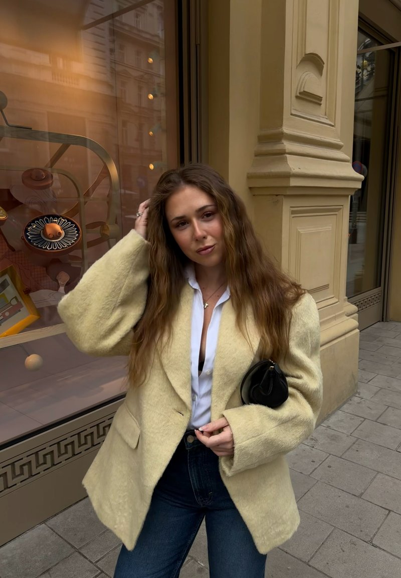 Jeune femme aux longs cheveux ondulés portant un manteau crème duveteux, une chemise blanche et un jean bleu, debout sur un trottoir en ville près d'une vitrine de magasin.