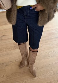 Άτομο που φορά σκούρο τζιν σορτς μέχρι το γόνατο, μπεζ καστόρινες μπότες με ψηλό τακούνι και καφέ παλτό από γούνα, στέκεται σε ξύλινο πάτωμα.