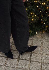 Personne portant un pantalon large noir et des bottes à talons pointues noires debout à côté d'un buisson vert décoré de guirlandes lumineuses jaunes.