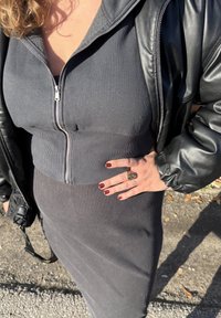 Vestito grigio a coste con corpetto aderente e chiusura con cerniera, sovrapposto a un giubbotto di pelle nera. Mano decorata con un anello e smalto scuro.