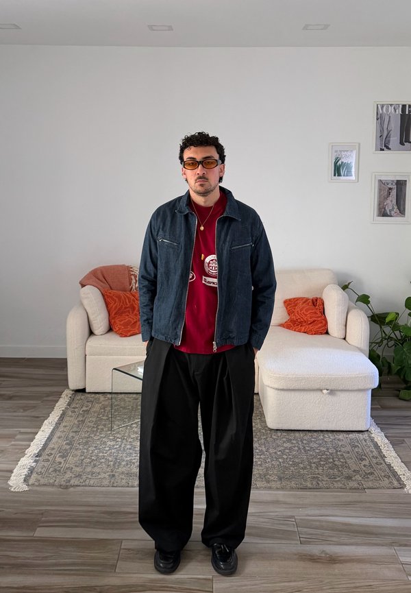 Mann mit oranger Sonnenbrille, rotem Hemd, dunkler Jacke und weiter schwarzer Hose steht in einem modernen Wohnzimmer mit weißem Sofa und orangenen Kissen.