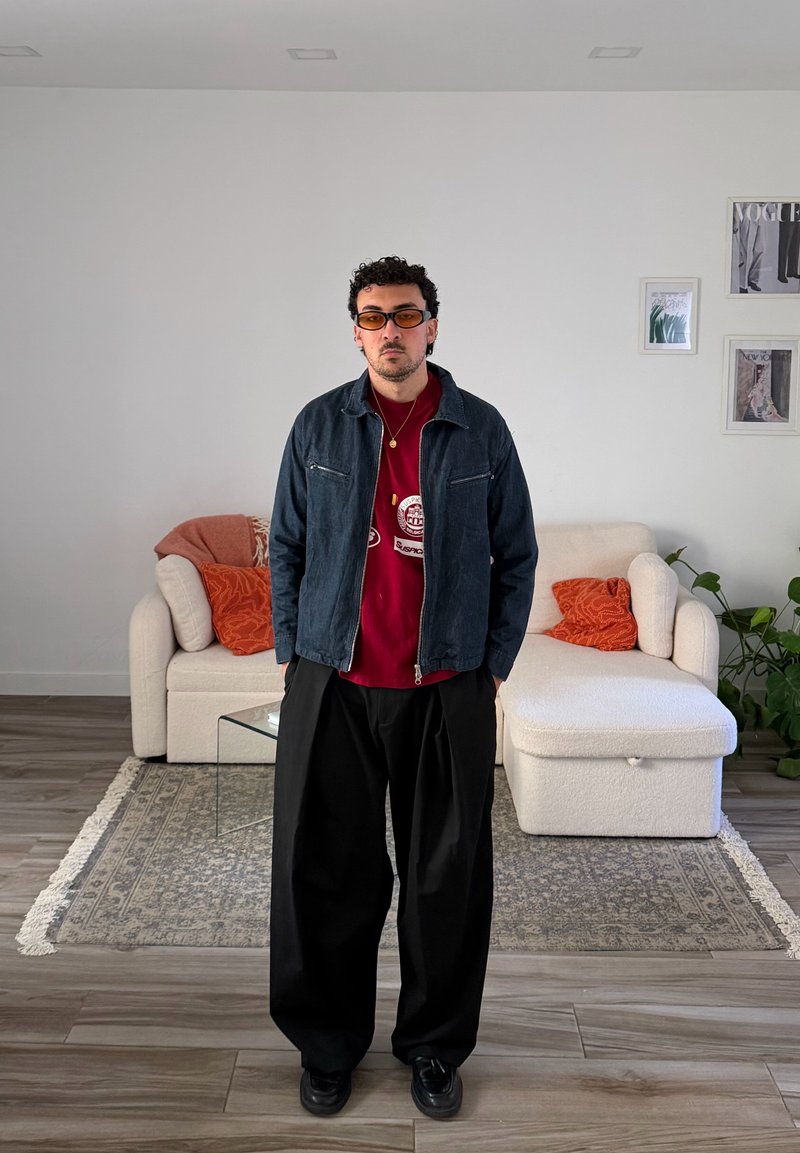 Homem com óculos de sol laranja, camisa vermelha, casaco escuro e calças pretas largas, está numa sala de estar moderna com sofá branco e almofadas laranja.