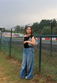 Μαύρη off-shoulder μπλούζα, φαρδιά μπλε τζιν και καρό τσάντα ώμου. Μακριά μαλλιά με χρυσά αξεσουάρ. Υπαίθριος χώρος με φράχτη.