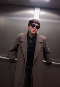 Person i en brun dubbelknäppt rock, bärande svarta handskar och mörka solglasögon, lutar sig avslappnat mot en metallisk hissvägg.