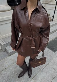 Bruine leren shirtjurk met knoopsluiting, een getailleerde taille en lange mouwen. Gecombineerd met bruine laarzen tot de knie en een kleine bruine handtas.