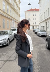 Prúžkovaná biela košeľa pod uhlovo pruhovaným sako, nosená s džínsami. Slnečné okuliare a dlhé tmavé vlasy viditeľné. Mestské prostredie.